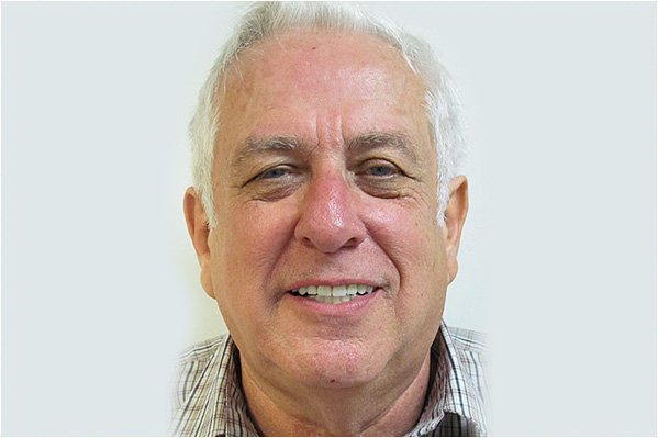 The image shows an older man with gray hair wearing glasses, smiling slightly, dressed in a patterned shirt, against a plain white background.