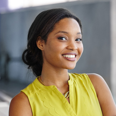 A smiling woman with dark hair wearing a yellow top and standing against a concrete wall.