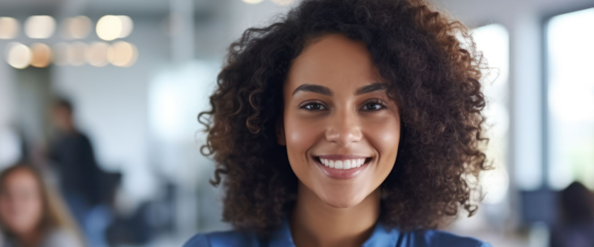 The image features a smiling woman with curly hair, wearing a blue top, standing indoors with blurred background elements suggesting an office environment.