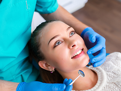 A dental hygienist performing a teeth cleaning procedure on a woman's mouth, with the woman sitting in a dental chair and holding her chin.