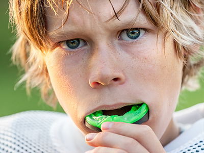 A young boy with blonde hair, wearing a football jersey, holding a mouthguard in his mouth.